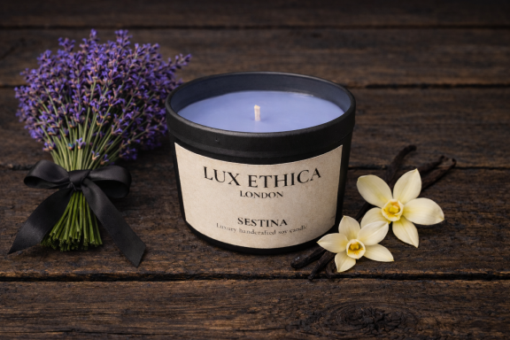 A lavender candle beside fresh lavender and two white flowers on a wooden surface.
